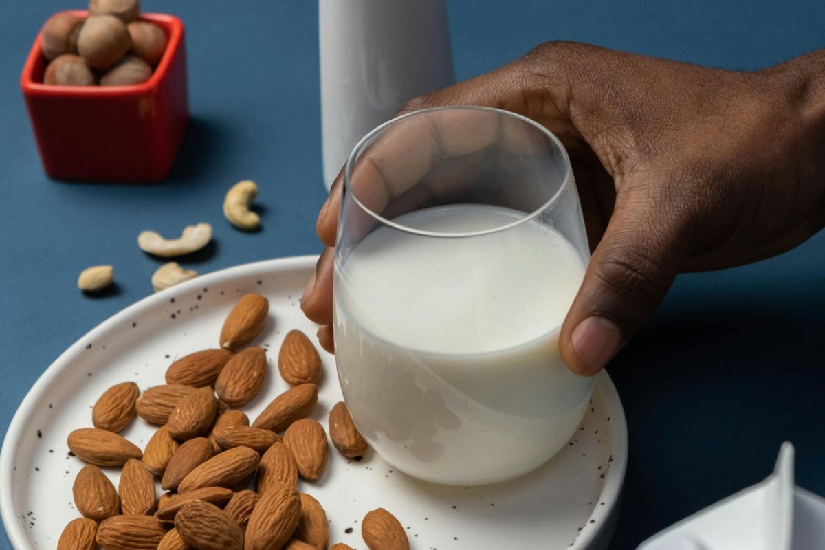 Uma mão segura um copo de leite ao lado de pratos com amêndoas e castanhas, mostrando alimentos que são boas fontes de cálcio e vitamina D.